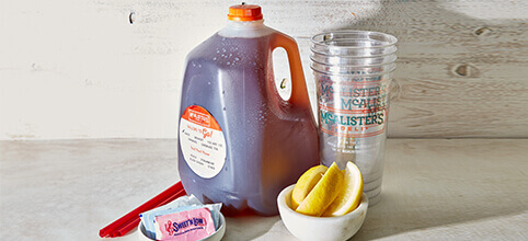 A gallon of McAlister's famous tea, with plastics cups, sliced lemon and packets of sugar on the side of it. 